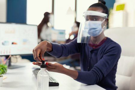 African entrepreneur using hand sanitizer at workplace wearing face mask. Businesswoman in new normal workplace disinfecting while colleagues working in background.