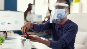 African entrepreneur using hand sanitizer at workplace wearing face mask. Businesswoman in new normal workplace disinfecting while colleagues working in background.