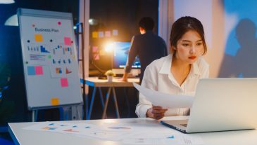 Asian businessman turn off computer and say goodbye his colleague who still working when he get off work after finish working overtime in small modern home office night. Coworker partnership concept.