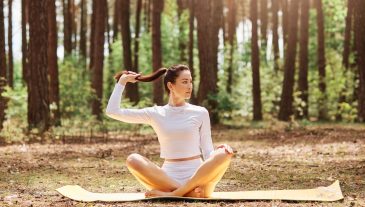 Attractive confident female wearing white top and leggins sitting on keremat on ground with crossed legs in lotus pose, looking thoughtfully aside, touching her ponytail.