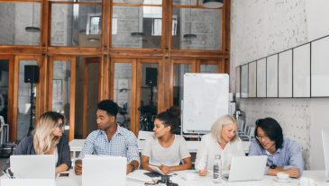 Blonde girl in white shirt talking with asian friend and drinking coffee near laptop with flipchart on background. Freelance web-designers working together in conference hall and using computers.