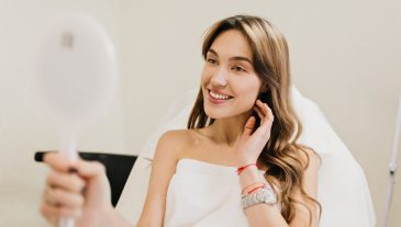 Happy beautiful woman with long brunette hair after cosmetology therapy smiling to mirror in white room. Joy, happines, good results, true positive emotions