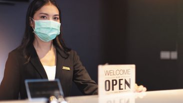 Business owner attractive young Asian woman in suitcase we're open sign on front counter welcoming clients to new hotel.Happy waiter with protective face mask holding open sign while stand at hotel .