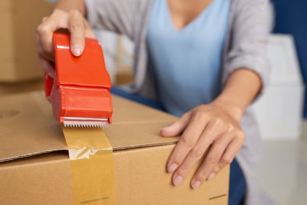 Close-up view of unrecognizable woman sealing cardboard box with adhesive tape while preparing for moving out