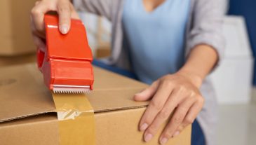 Close-up view of unrecognizable woman sealing cardboard box with adhesive tape while preparing for moving out